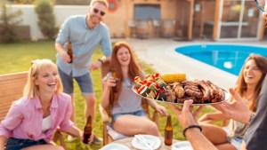 A group of friends enjoying a barbecue by a pool, with one person serving a platter of grilled meats and vegetables while others smile and hold drinks.