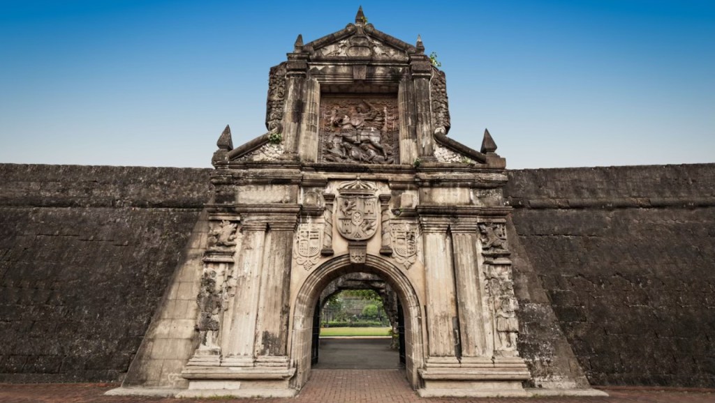 A historic entrance gate of Intramuros, Manila, featuring intricate sculptures and surrounded by stone walls under a blue sky.