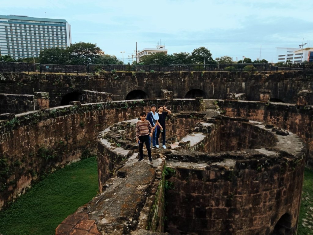 Three individuals standing on the ruins of a historical fortification, surrounded by lush greenery and modern buildings in the background, showcasing the blend of history and contemporary urban life.