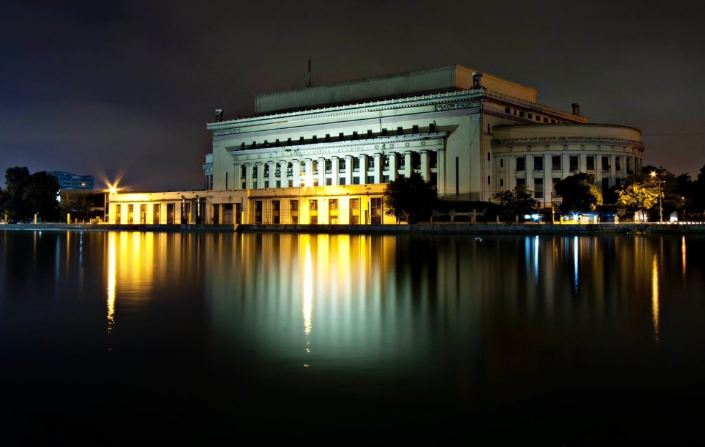 A nighttime view of a grand building reflecting in calm waters, illuminated by golden lights.