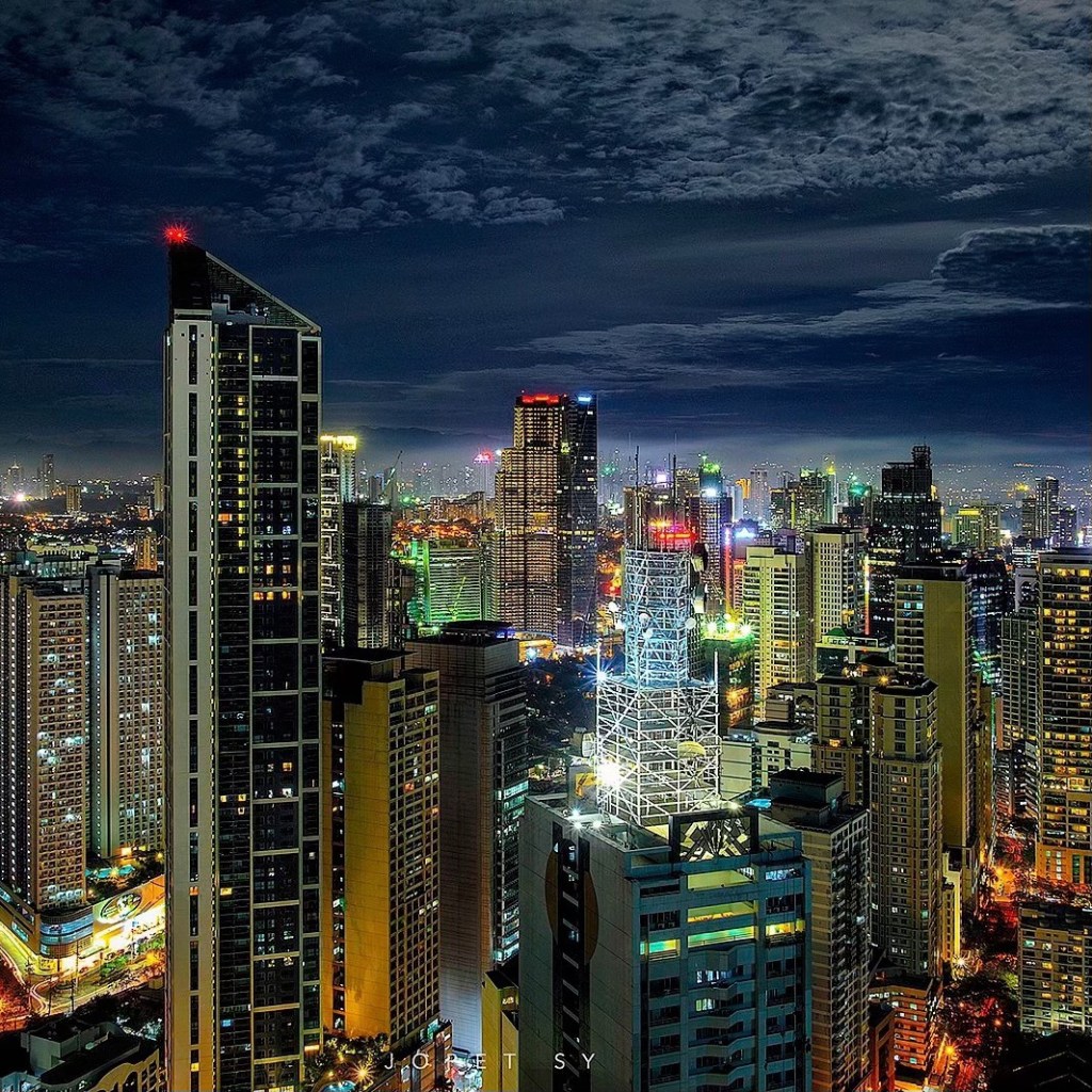 Night view of a vibrant city skyline with illuminated skyscrapers and a cloudy sky.