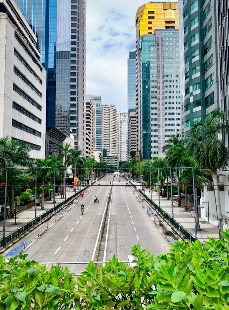 A wide view of a central business district street lined with tall buildings and palm trees, showing a mostly empty road with few pedestrians and cyclists.