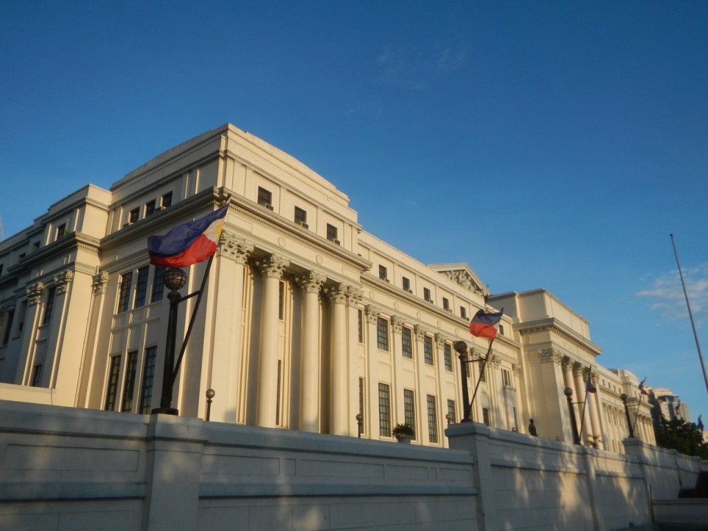 A grand colonial building in the Philippines featuring neoclassical architecture, adorned with flags and set against a clear blue sky.