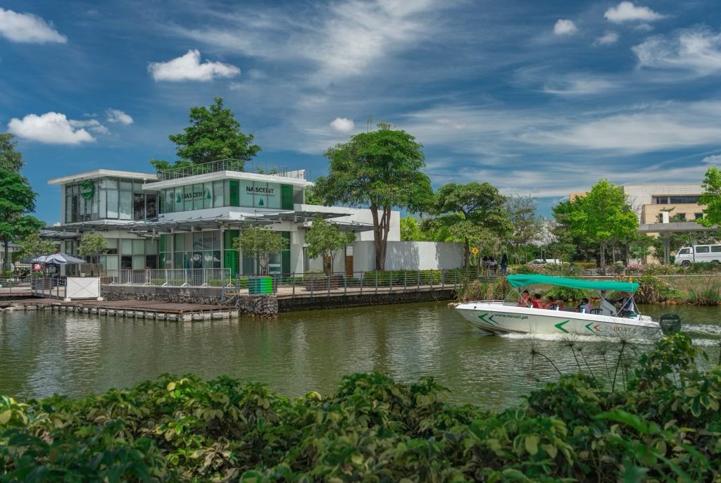 A scenic view of a modern building next to a tranquil lake, with a boat moving across the water and lush greenery surrounding the area.