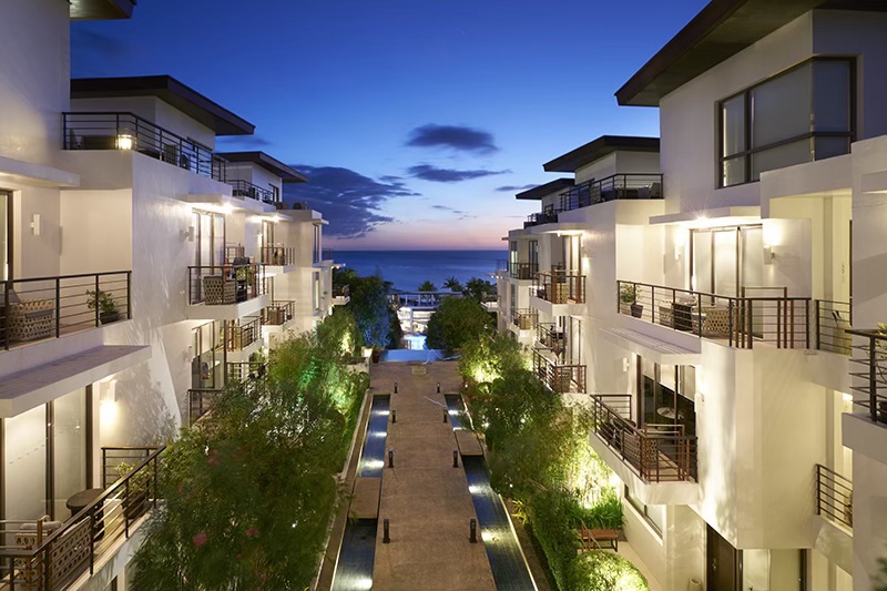 A luxurious beachfront property in the Philippines at dusk, showcasing modern architecture with balconies and a pathway lined with greenery leading to the ocean.