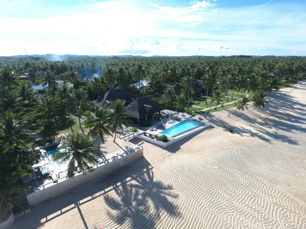 Aerial view of a luxurious beachfront property in the Philippines with a swimming pool, surrounded by lush palm trees and sandy beach.