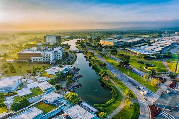Aerial view of a modern urban township with lush greenery, a winding waterway, and various commercial buildings under a blue sky during sunset.