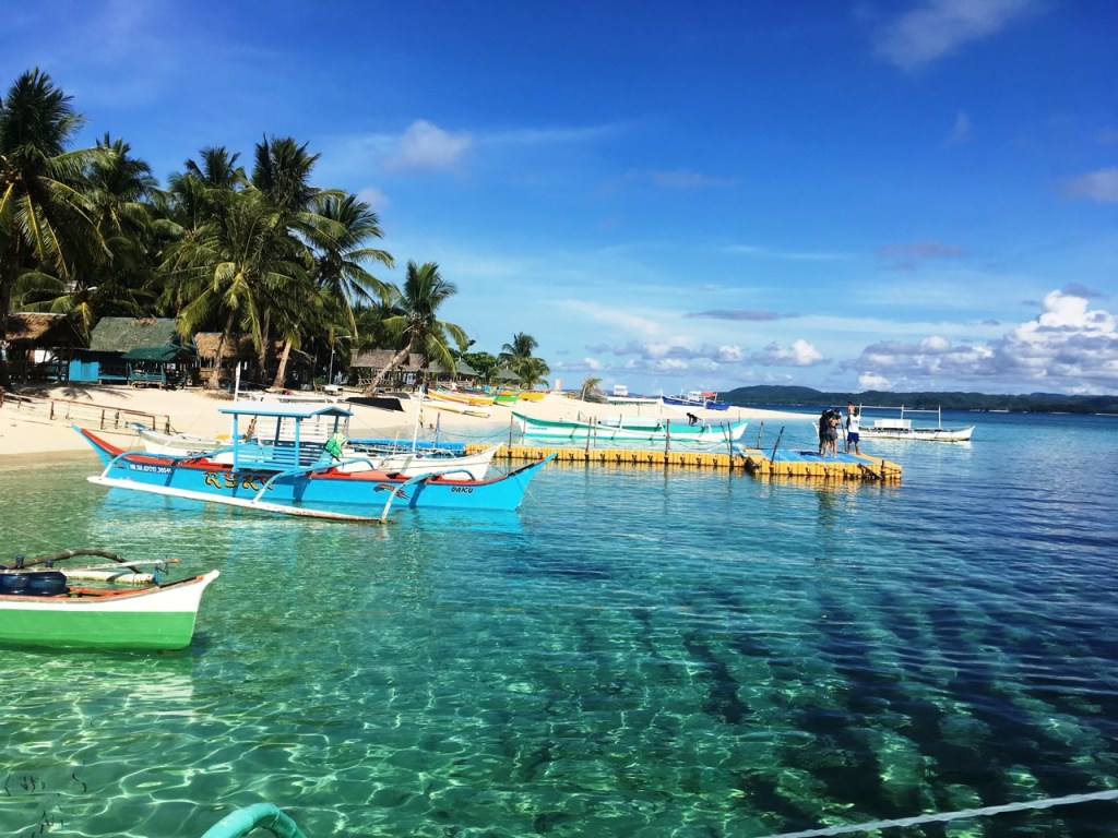 A picturesque beachfront scene in the Philippines, featuring clear turquoise waters, traditional banca boats moored at a wooden jetty, and palm trees lining the sandy shore under a bright blue sky.