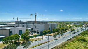 Construction site with cranes and modern buildings alongside a wide road, set against a clear blue sky.
