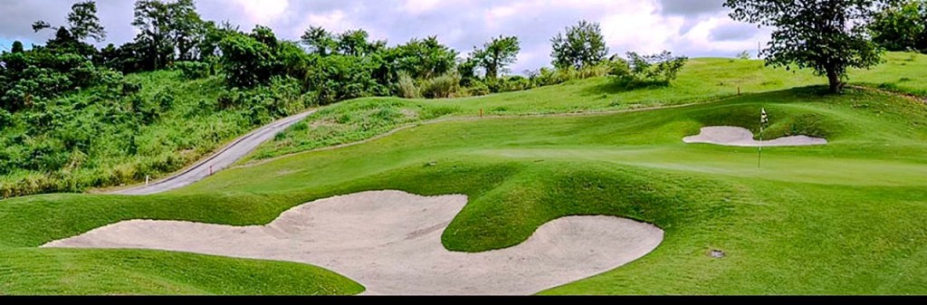 A scenic view of a golf course in Silang, Cavite, featuring lush green fairways, sand traps, and a winding path amidst rolling hills and trees.