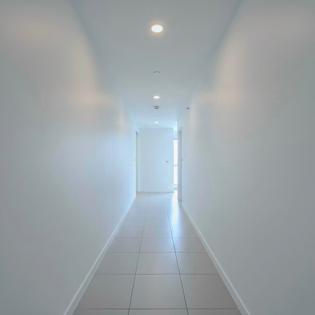 A long, empty hallway with white walls and tiled flooring, featuring bright ceiling lights and a door at the far end.