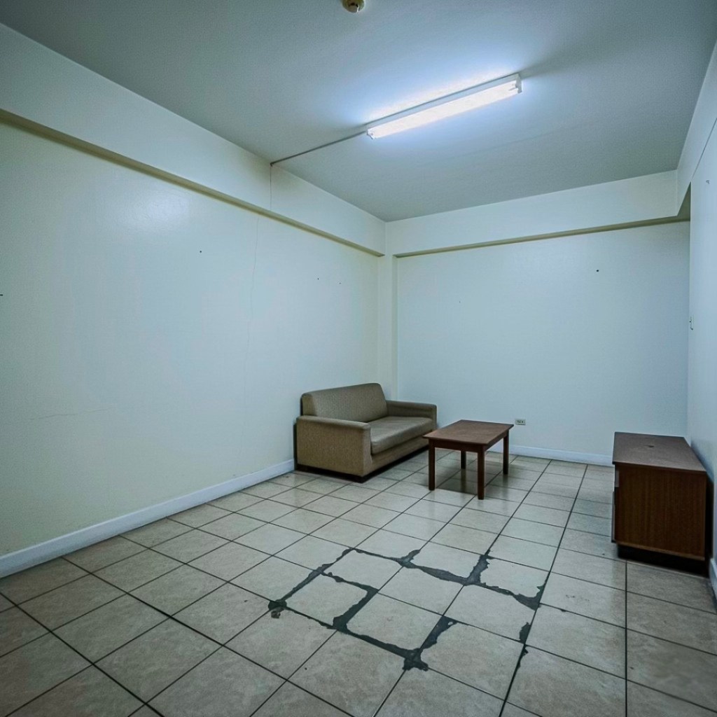 An empty condo living space featuring a worn-out tiled floor, a simple sofa, and a wooden coffee table, with plain white walls and overhead fluorescent lighting.