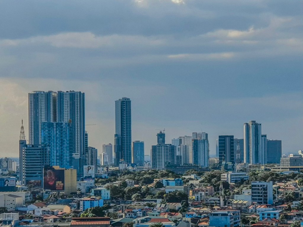 A panoramic view of a modern city skyline featuring tall skyscrapers and a mix of residential buildings beneath a cloudy sky.