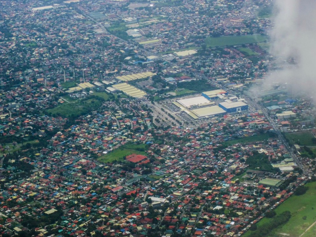 Aerial view of a densely populated urban area, featuring a mix of residential buildings and commercial structures, with green spaces interspersed throughout.