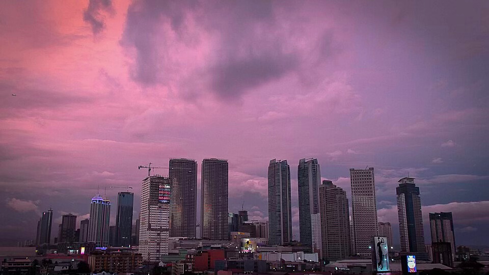 City skyline during sunset with pink and purple clouds, featuring tall skyscrapers and some illuminated buildings.