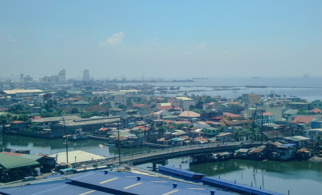 A panoramic view of a coastal city featuring residential buildings, a river, and industrial structures, with ships in the distance under a clear blue sky.