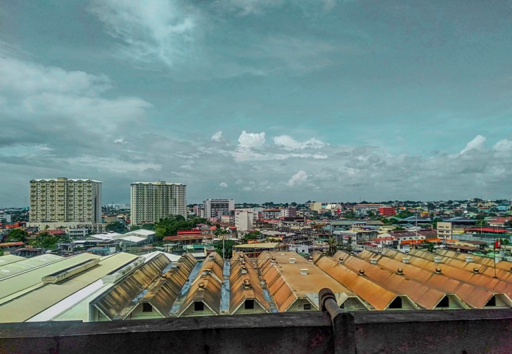 Panoramic view of a cityscape featuring a mix of residential and commercial buildings, with a cloudy sky above.