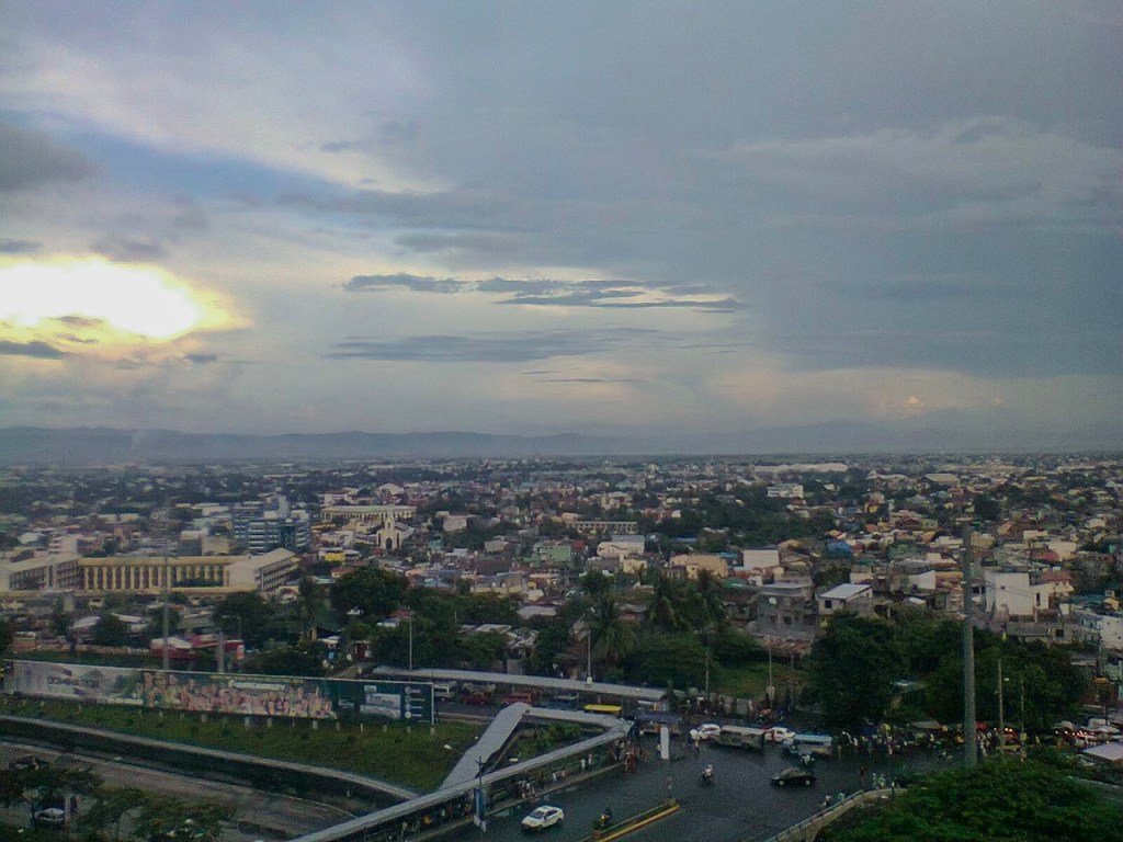 A panoramic view of a cityscape under a cloudy sky, featuring a blend of residential and commercial buildings with distant mountains in the background.