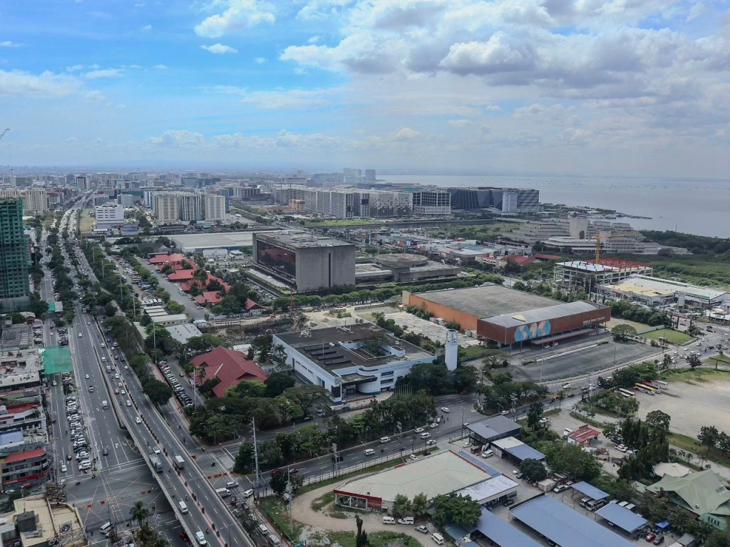 Aerial view of a cityscape with a mix of urban buildings and greenery, featuring roads with traffic, large commercial structures, and a body of water in the background under a partly cloudy sky.